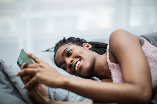 Young adult back woman lying down in bed with mobile and chatting