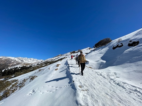 uttarakhand, India – December 12, 2023: Group of people ascending a snowy mountain slope with a bright blue sky in the background