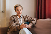 Medium shot of middle-aged woman resting on sofa , using applications on smartphone in living room. Female chatting in messenger or social network with friends