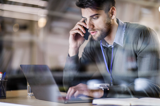 Young male entrepreneur talking on cell phone while working on a computer in the office. The view is through glass.