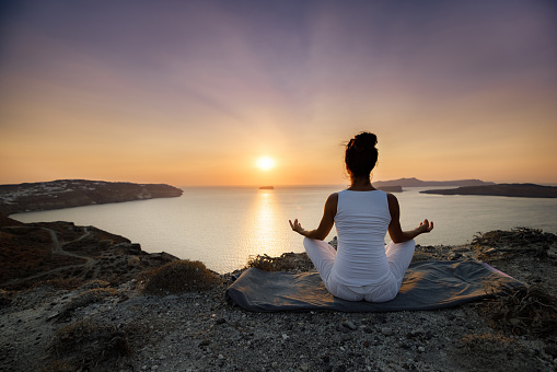 Back view of a relaxed woman doing Yoga meditation exercises in Lotus position on a hill above the sea at sunrise. Copy space.
