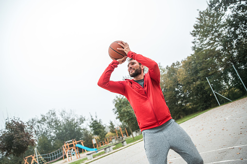 A Hispanic male, wearing a red hoodie, takes a shot while playing basketball outdoors.