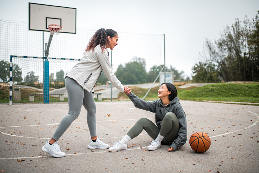 A young adult Hispanic female friend assists her Asian friend after a fall during their outdoor basketball game, showcasing camaraderie among diverse friends.