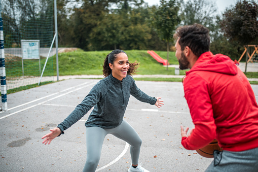 A joyful Hispanic woman in athletic gear prepares to defend against a Caucasian man in a red jacket during an outdoor basketball game.