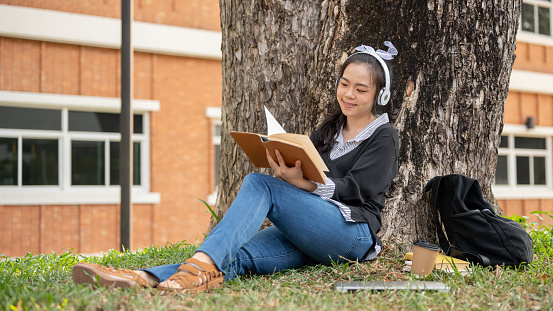 A pretty and positive young Asian female college student in casual outfit is listening to music on her headphones and reading a book, relaxing under the tree in a campus park. Lifestyle and leisure