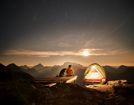 Rear view of a man with his pet dog on vacation sitting on wooden deck on the edge of mountain next to the tent in the evening and admiring the sunset and night starry sky