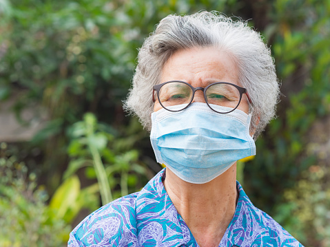 A portrait of an elderly Asian woman wearing a face mask looking at the camera while standing in a garden. Mask for protect virus, coronavirus, pollen grains. Concept of old people and healthcare.