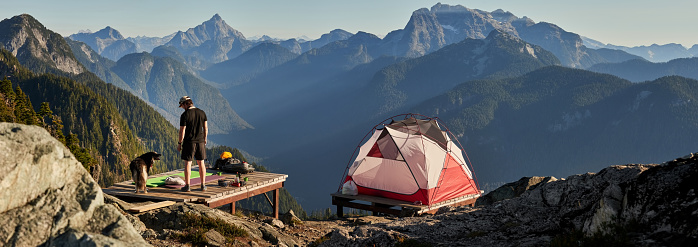 Rear view of a young man with pet dog standing on wooden camping deck on mountain top with tent on side
