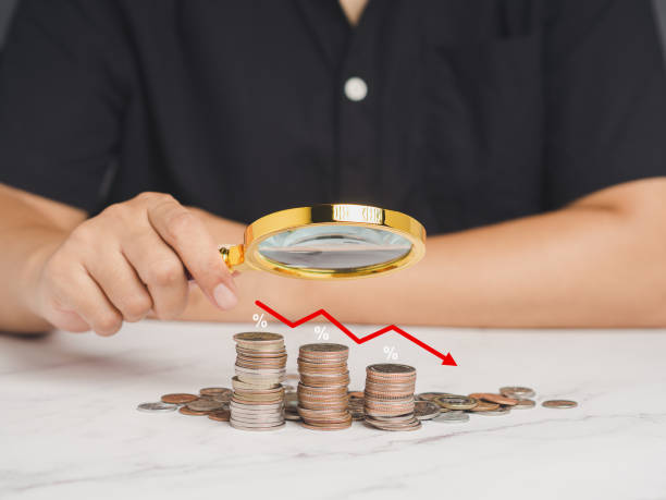 istock Hand holding a magnifier looking at stacked coins on a table with an interest rate decreasing graph. 1854258093