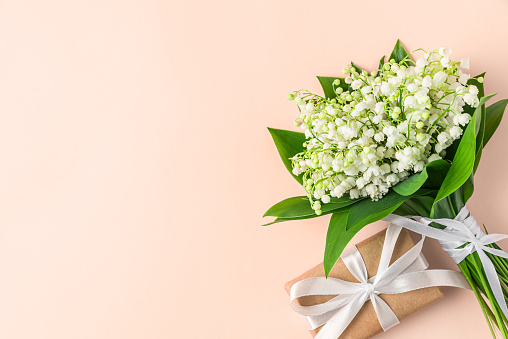 Lily of the valley flowers bouquet with gift box on peach color background. Womens day, Valentines day card. Top view with copy space. Flat lay. Trendy color 2024.