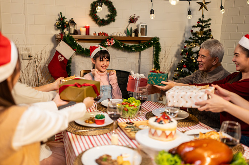 Asian family giving gift box to young daughter during Christmas party. Attractive little girl receive presents from relative while having dinner celebrate holiday Thanksgiving on dining table together