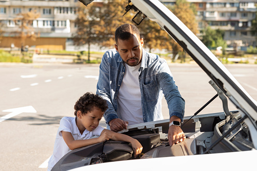 Upset concerned black dad and child standing on parking slot, fixing engine in broken automobile. Young african american father teaching his little son to repair some parts in family car