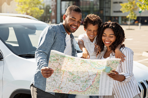 Loving african american family father mother and preteen son going car trip together, standing on the street next to white automobile, checking map, looking for destination to travel