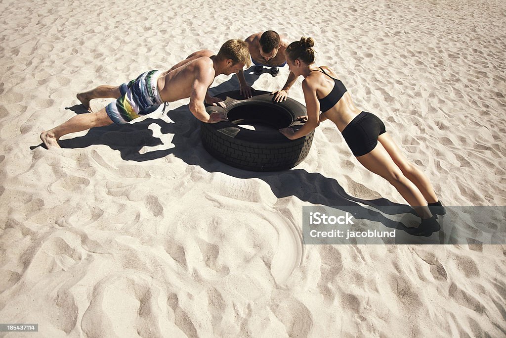 Young athletes doing push-ups on tire Small group of people doing push-ups on tire. Young athletes working out on beach Cross Training Stock Photo