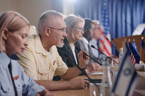 Mid adult Caucasian man holding microphone is asking questions during a community town hall meeting or debate. Candidate or speaker is in background standing at podium during speech.