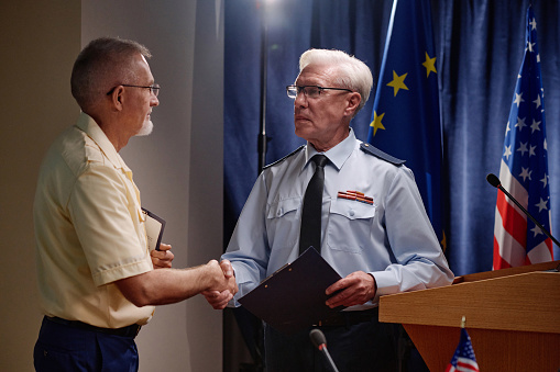 Vertical photo of mature man and woman in business suits standing in hallway of building and shaking hands while looking at each other