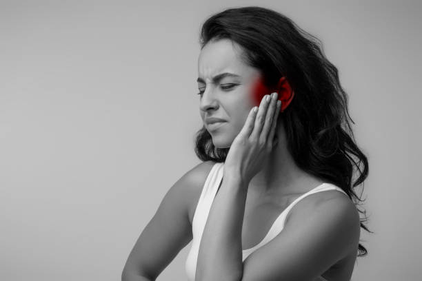 Young brunette eastern lady suffering from otitis, touching ear Black and white photo of young brunette eastern lady suffering from otitis, rubbing her inflamed ear highlighted with red over grey background, ear diseases concept, copy space adult ear pain stock pictures, royalty-free photos & images