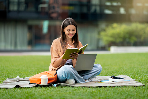 Cheerful beautiful young woman in stylish casual outfit university student working on project, sitting on lawn at city park, using laptop computer outdoors, drink takeaway coffee, reading book