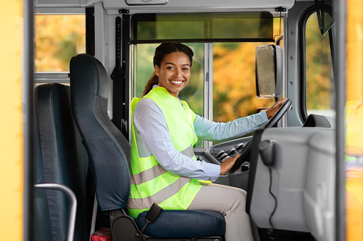 Happy black female driver sitting inside of bus and looking at camera, smiling beautiful african american woman in reflecting clothing holding steering wheel, waiting passengers, enjoying her work