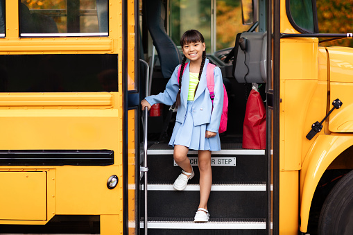 Smiling preteen asian girl getting of the yellow school bus, happy female kid wearing backpack stepping down off of the vehicle as she arrives for study, ready for lessons and learning fun
