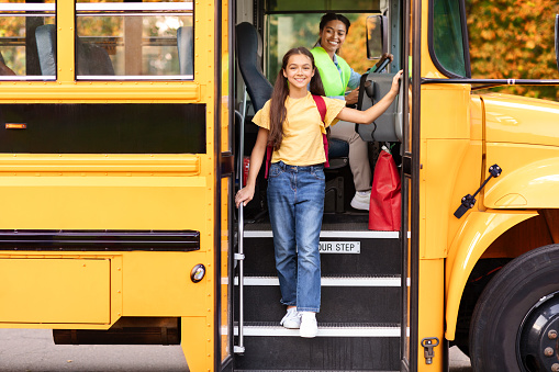 Happy preteen girl getting of the yellow school bus, cheerful female kid stepping down off of the vehicle, friendly black female driver in uniform looking at her and smiling, free space