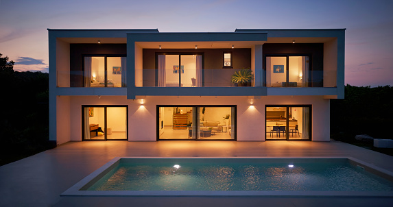 Exterior view of illuminated modern luxury house with swimming pool in foreground at dusk.