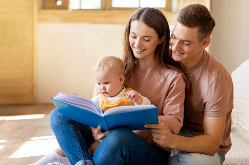 Happy Family Of Three With Cute Infant Son Reading Book Together At Home, Young Loving Parents Spending Time With Their Adorable Toddler Kid, Relaxing On Floor In Living Room, Copy Space