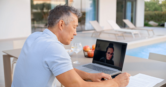Businessman and businesswoman using laptop for online meeting in video call while sitting by swimming pool at home.
