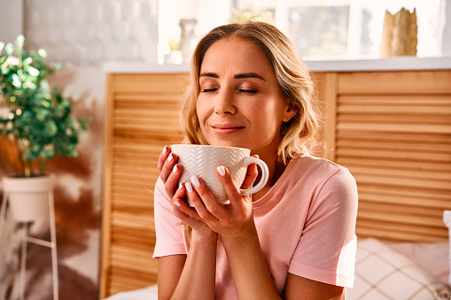 Portrait of pleasant woman with wavy blond hair holding white coffee cup with two hands and closing eyes from enjoyment. Relaxed young lady wearing comfy pink pajama in bed.