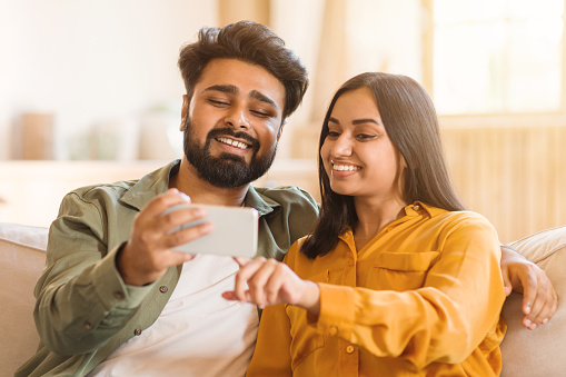 Young Indian couple sitting comfortably indoors, sharing joyful moment as they take selfie together. Both exhibit genuine smiles, capturing a candid and delightful shared experience