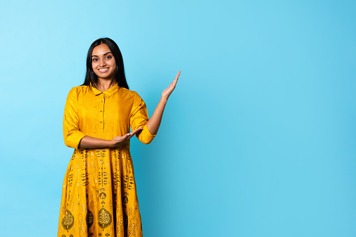 Fashion Offer. Beautiful Indian Young Woman Wearing Traditional Casual Yellow Dress With Golden Embroidery, Posing Showing Empty Space For Text Standing Over Blue Background. Studio Shot