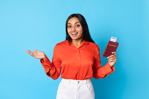Travel Tour Offer. Excited Middle Eastern Tourist Woman Holding Boarding Pass And Passport Standing Over Blue Background, Glad About New Vacation Journey, Advertising Cheap Flight Tickets