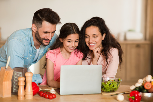 Happy european family of three using laptop, browsing healthy recipes while preparing dinner together, kitchen interior. Parents and daughter cooking