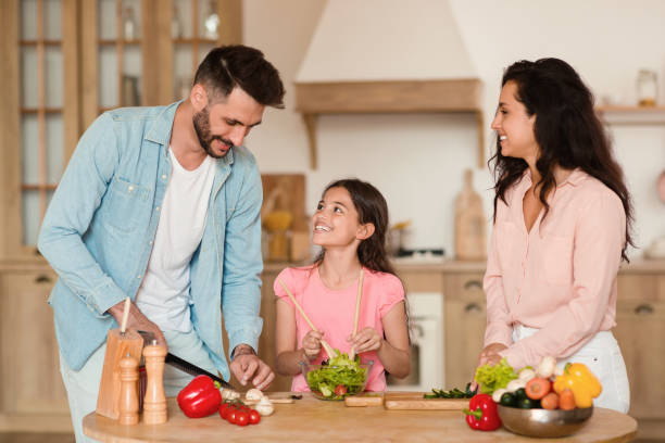 loving parents cooking with their cute daughter, mother and father cutting vegetables while girl mixing fresh salad - two parent family indoors home interior domestic kitchen imagens e fotografias de stock