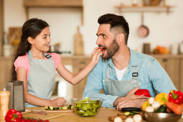 menina alimenta pai com fatia de tomate enquanto cozinham juntos - healthy eating snack child domestic kitchen - fotografias e filmes do acervo