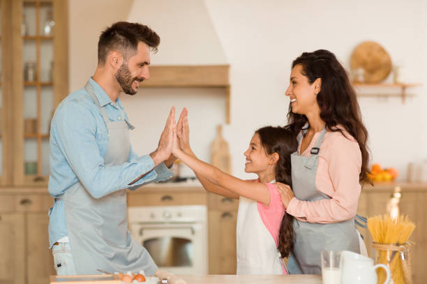 cute girl and her dad giving high five to each other while cooking in kitchen, loving parents and daughter having fun - two parent family indoors home interior domestic kitchen imagens e fotografias de stock