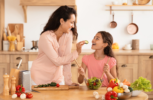 Cheerful Young Mother And Daughter Having Fun While Cooking Together, Feeding Kid Girl Giving To Taste Cucumber Slice In Modern Kitchen Indoors. Tasty And Healthy Family Nutrition