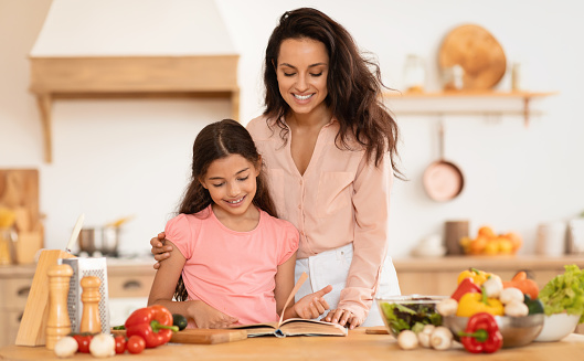Family Dinner Preparation. Cheerful Mom And Kid Daughter Cooking And Reading Recipes Book In Modern Kitchen Indoor, Standing Near Table Making Healthy Fresh Salad Together