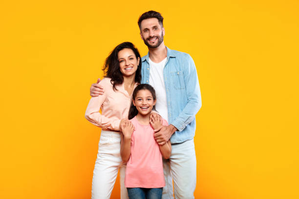 Happy european family of three hugging and smiling to camera, posing together over yellow studio background stock photo