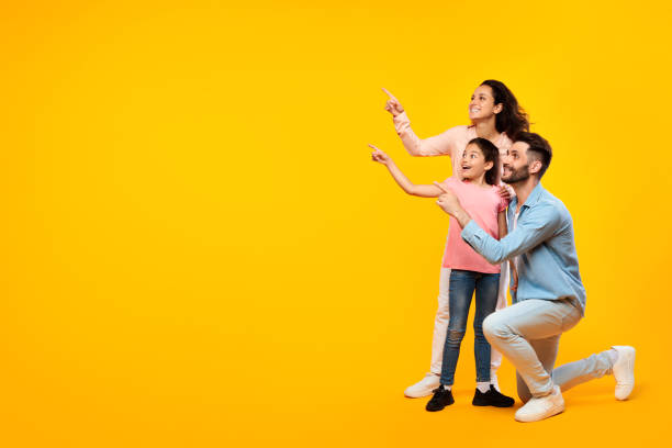 istock Excited family of three pointing aside at free space while posing over yellow studio background, full length, banner 1854549318