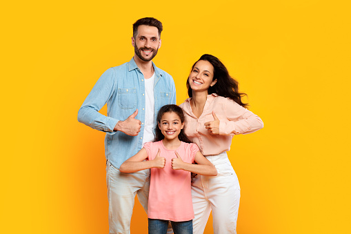 We like it. Portrait of smiling european family of three showing thumbs up gesture, approving or recommending something, standing on yellow background