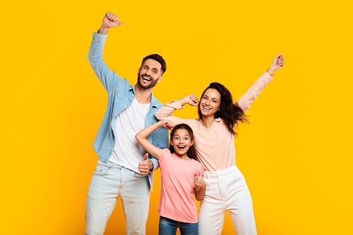 Victory concept. Overjoyed european family of three shaking clenched fists, making winner gesture, posing over yellow background. Father, mother and their daughter celebrating success together