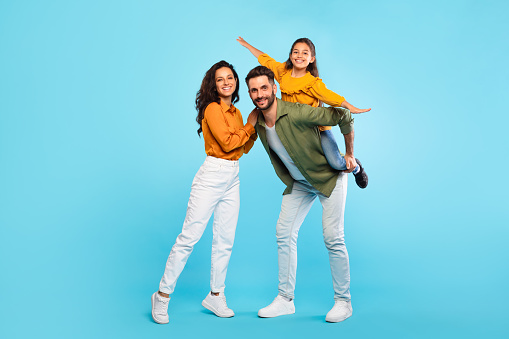 Cheerful european family of three having fun, dad piggybacking daughter over blue background, parents and their child smiling at camera, full length, free space