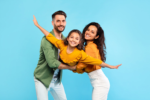 Portrait of happy european parents fooling with their daughter, holding child on hands and girl pretending flying, spreading hands, having fun over blue background