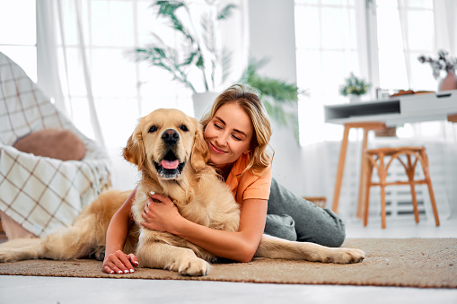 Adorable woman with closed eyes embracing adult golden retriever while lying together on floor.