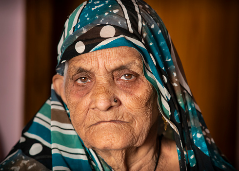 Indoor close-up portrait of a senior Indian woman looking at the camera with a blank expression. She is wearing a sari and covering the head.