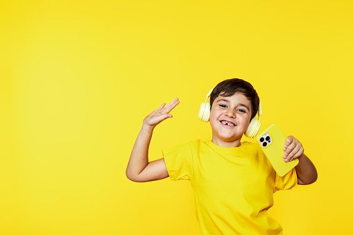 Cheerful Caucasian boy with headphones holds a phone, enjoying tunes on a bright yellow background.