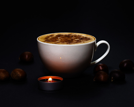 A white cup of coffee topped with cinnamon, surrounded by chestnuts and a small perfumed burning candle, during winter and holiday season, bright and dark contrast and chiaroscuro, fire light reflection on the mug