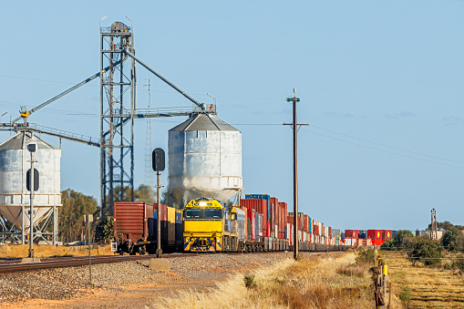 Huge grain storage tanks provide an imposing background to a double-stack container freight train as it passes another container train in a rural industrial location. The front of the northbound train is almost parallel with the rear of the southbound train with its electronic end-of-train device. A healthy load of double stack containers is visible. In the foreground, are the railway signals for the passing siding (loop).