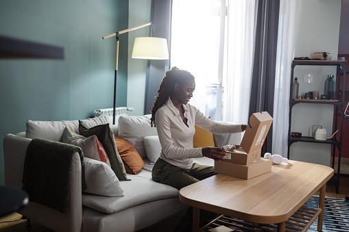 A young African American woman sitting in her cozy living room, while cheerfully opening her new package that has just arrived.